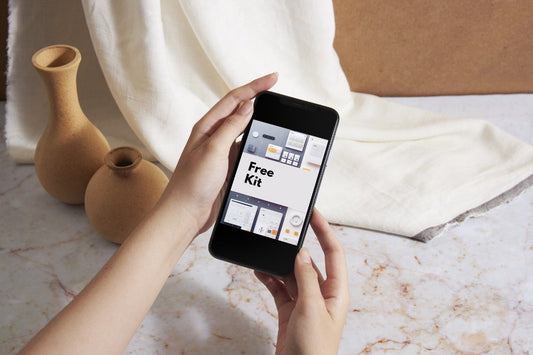 A person holds a black smartphone showing a mockup screen with the text 'Free Kit' over a styled tabletop scene with beige vases, draped cream fabric, and a marble surface.