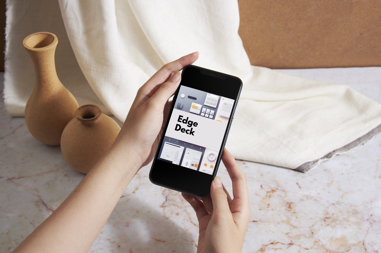 A person holds a black smartphone displaying the Edge Deck app interface, with a cream cloth and two small tan vases on a light marble surface in the background.