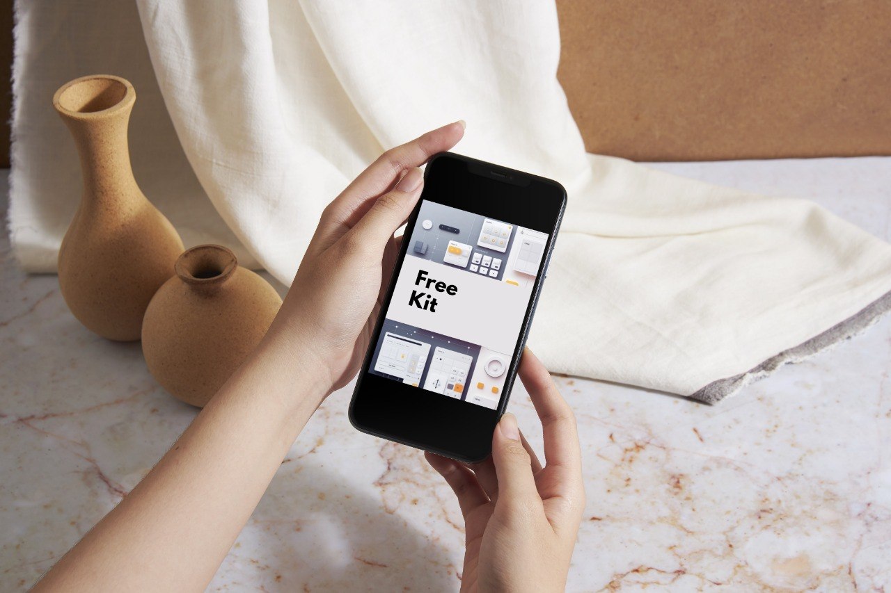 A person holds a black smartphone showing a mockup screen with the text 'Free Kit' over a styled tabletop scene with beige vases, draped cream fabric, and a marble surface.