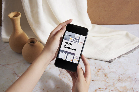 A person holds a smartphone showing the Shift Capsule app interface, with two terracotta vases and a draped cream fabric in the background on a marble surface.