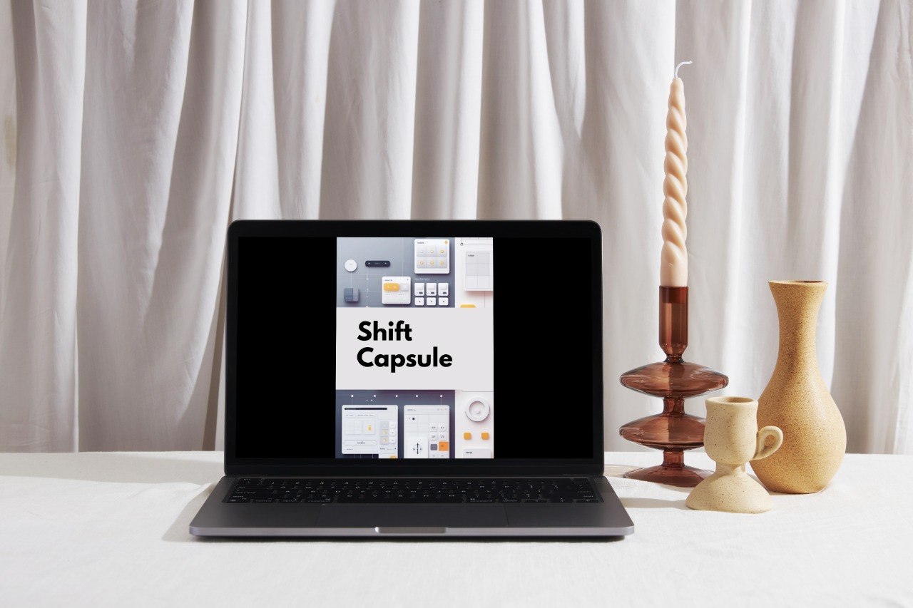 A black laptop sits open on a white surface, displaying a digital interface with the text 'Shift Capsule' on the screen. The setup is styled with decorative objects, including a tall spiral candle in a wooden holder, a small ceramic cup, and a beige vase, against a backdrop of draped white curtains.