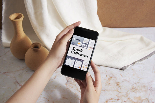 A person holds a smartphone showing the 'Spark Collection' app interface, with a cream fabric drape and two small terracotta vases in the background on a marble surface.